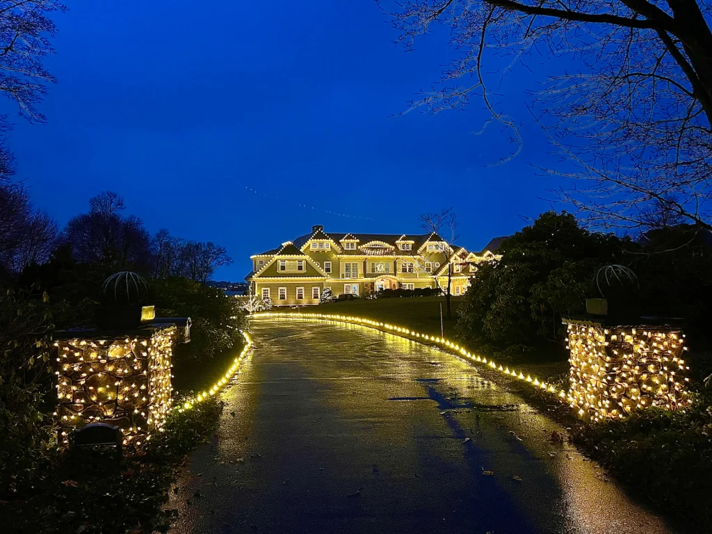Warm white holiday lighting on a large home, stone entry pillars, and a lit driveway at night