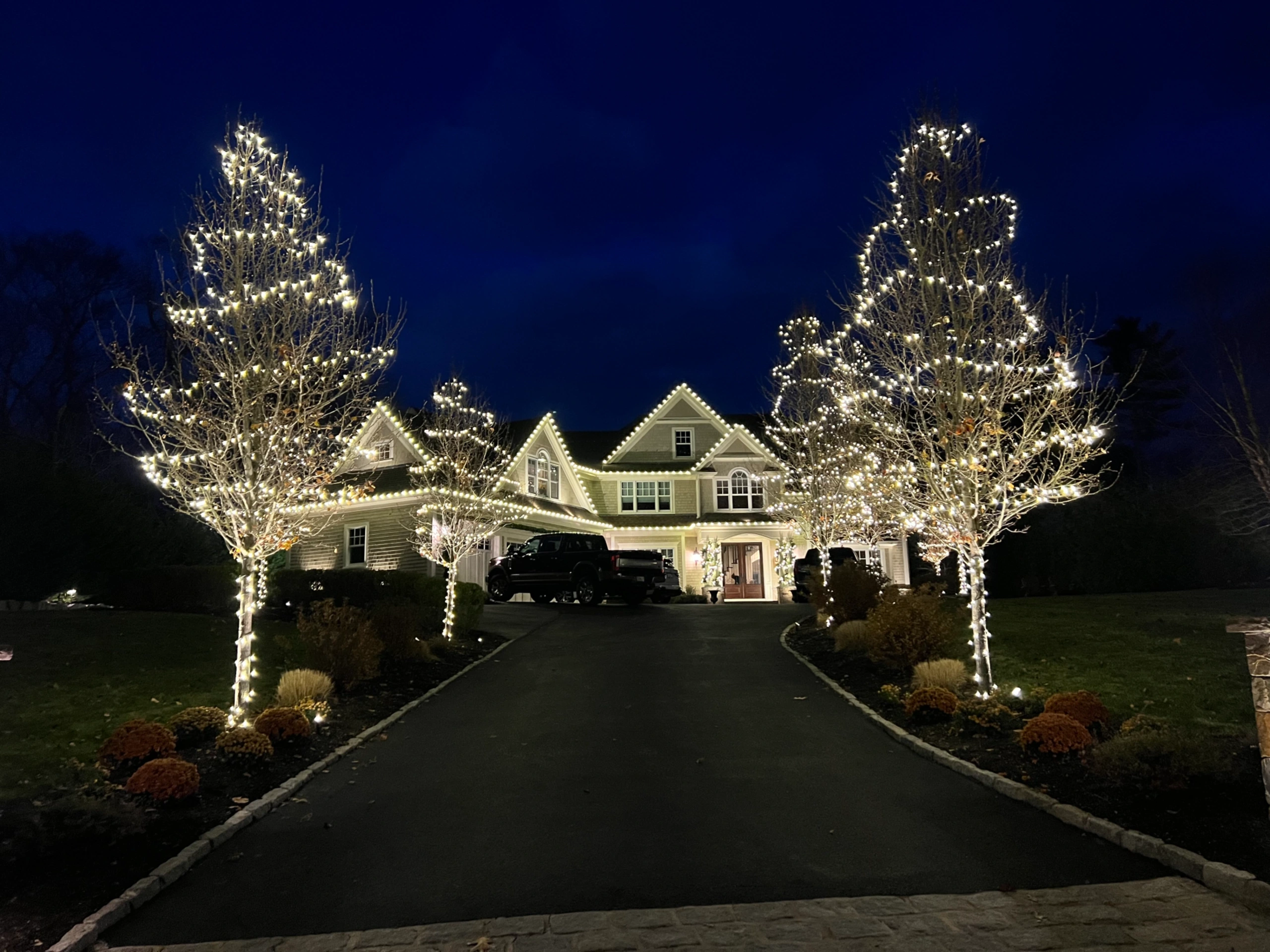 Warm white holiday lighting on a home's rooflines and driveway trees at night